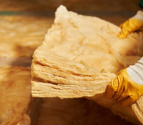 Main tenant un rouleau de laine minérale isolante, matériau d'isolation thermique beige et fibreux, avec des gants de travail jaunes.