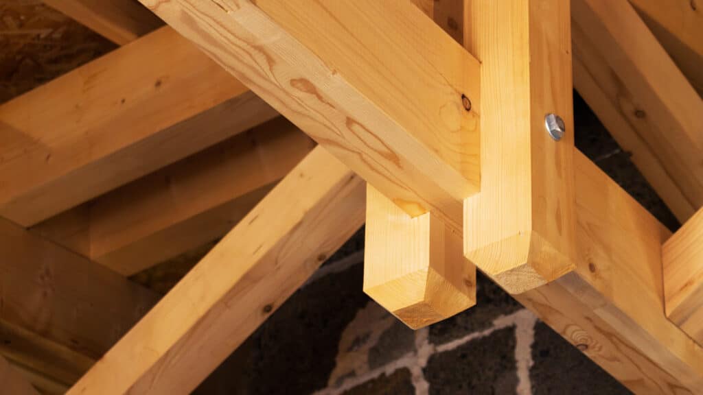 Assemblage robuste de charpente en bois clair, avec tenon et mortaise sécurisés par un boulon, au-dessus d'un mur en pierre sombre.