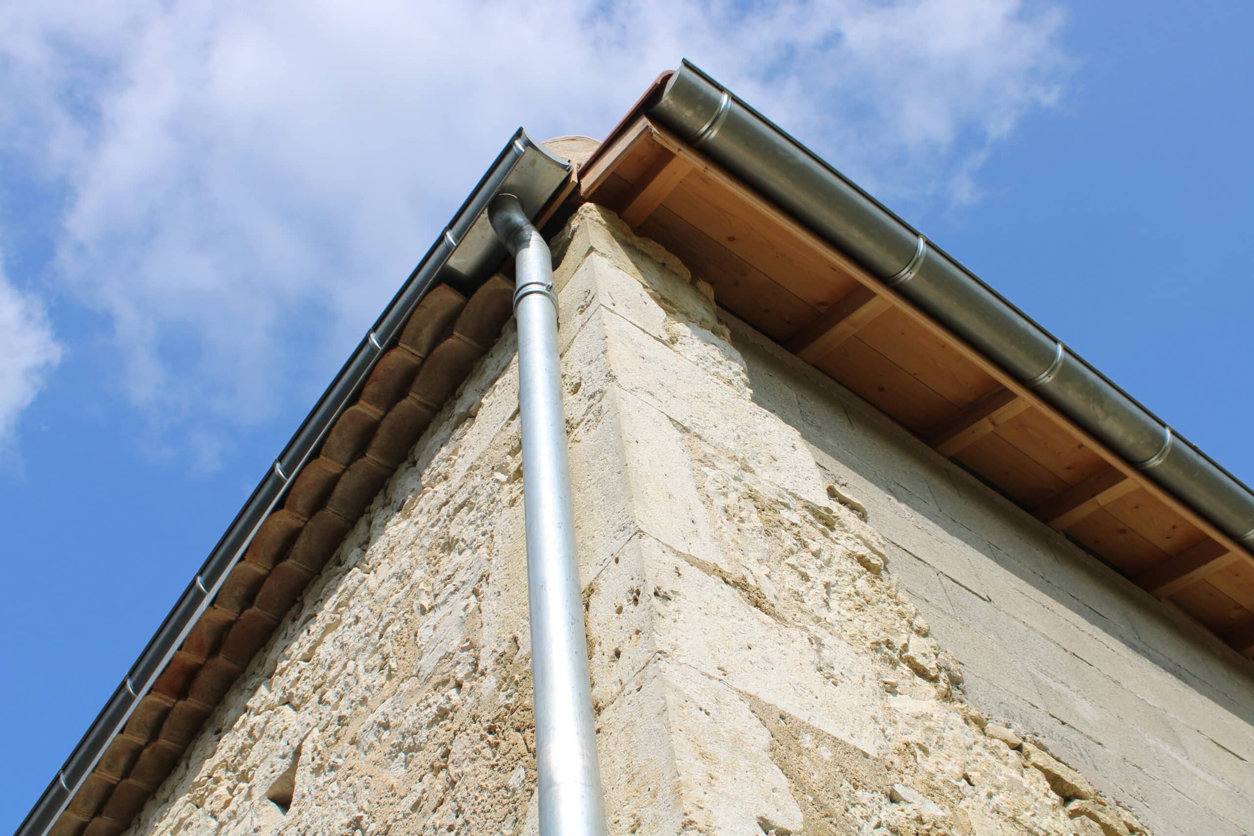 Gouttière zinc neuve et descente d'eau pluviale sur coin de mur ancien en pierre calcaire sous ciel bleu.
