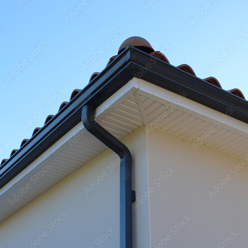 Gouttière noire et descente pluviale sur une maison avec soffite blanc et tuiles de toit en terre cuite, sous un ciel bleu clair.