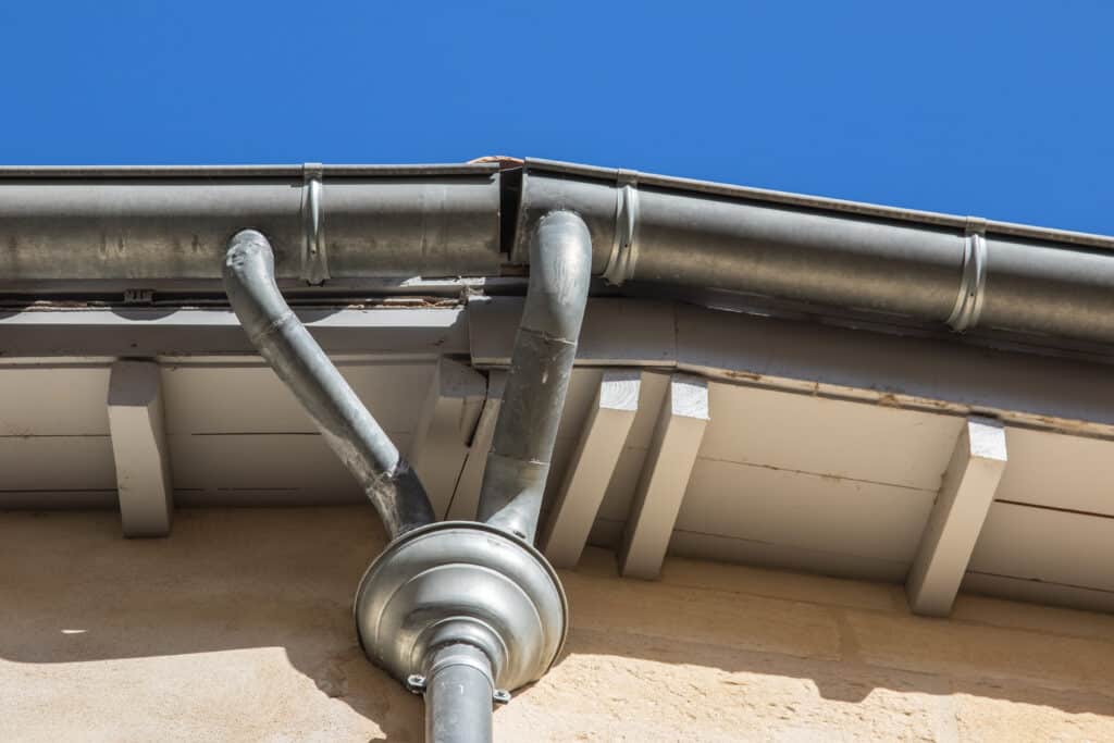Gouttière zinc et descente eau pluviale façade Gouttière en zinc sur façade crépie, avec descente d'eau pluviale et raccords coudés sous ciel bleu dégagé.