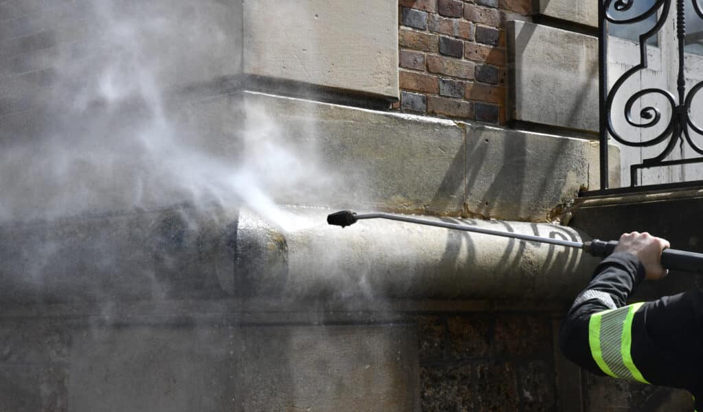 Nettoyage façade pierre : nettoyeur haute pression en action Un ouvrier en vêtement de sécurité jaune et noir nettoie une façade en pierre avec un nettoyeur haute pression, créant un panache de vapeur.