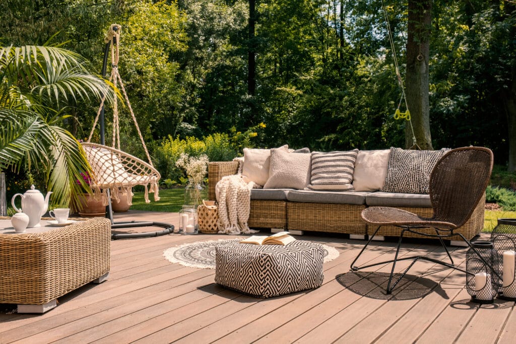 Salon de jardin bohème : détente sur terrasse bois Terrasse en bois aménagée avec mobilier en rotin, coussins beiges, pouf à motif géométrique et fauteuil suspendu près d'un jardin luxuriant.