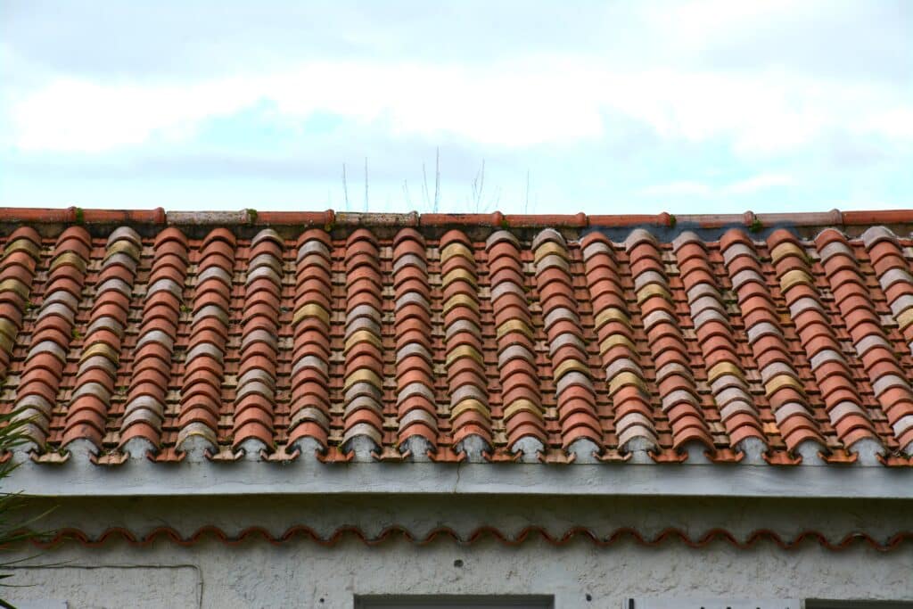 Toit en tuiles romanes rouges et usées Tuiles romanes rouges et grises vieillies sur le toit d'un bâtiment blanc, avec un ciel clair et nuageux au-dessus.
