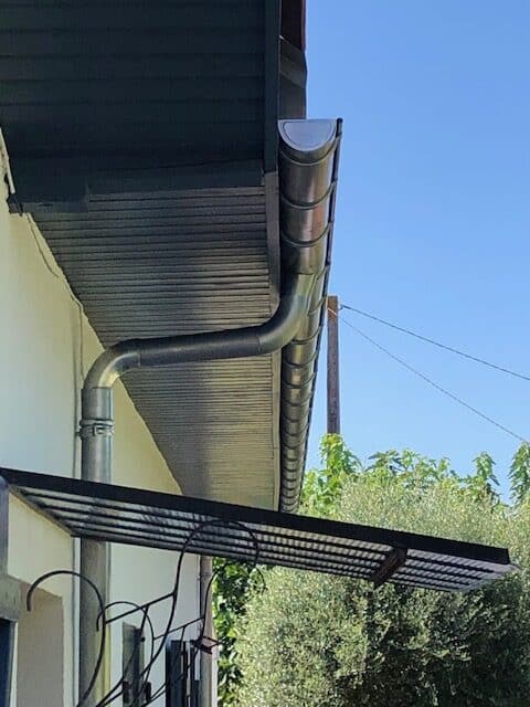 Système de gouttière en zinc avec descente d'eaux pluviales Gouttière en zinc avec descente d'eau pluviale et coude sur une façade sous un avant-toit en bois sombre.