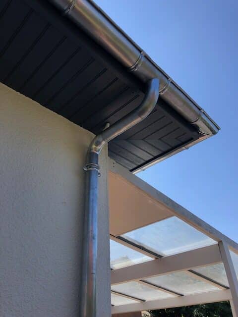 Installation de Gouttière et Descente d'Eau Pluviale Gouttière et descente d'eau pluviale en métal sur une façade de maison avec sous-toiture en bois sombre.