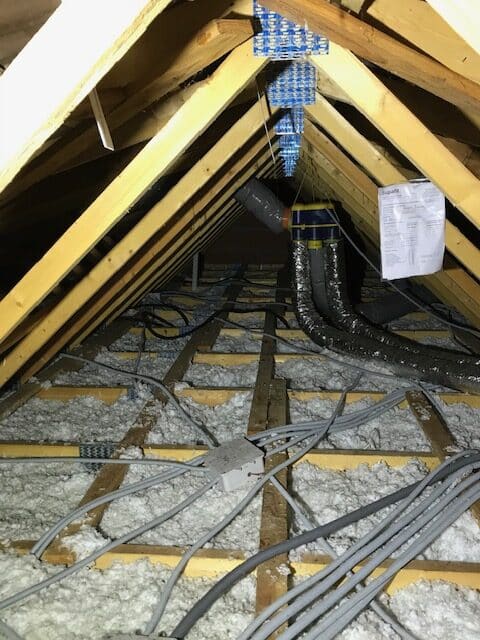 Combles avec charpente bois, isolation soufflée, gaines électriques grises et système de ventilation mécanique.